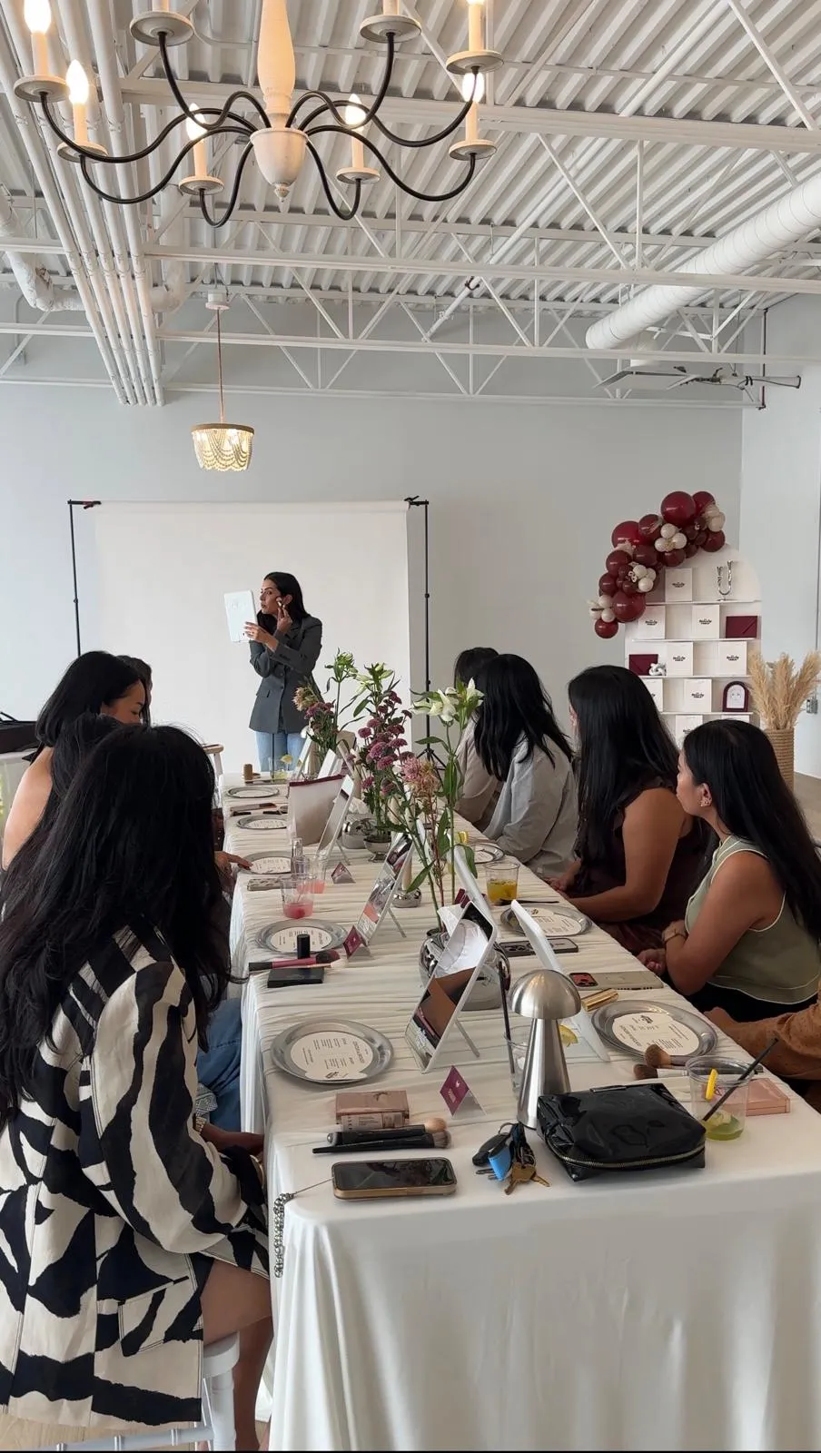 A host demonstrates a technique with a brush and mirror at the head of a long table while guests follow along from their place settings.