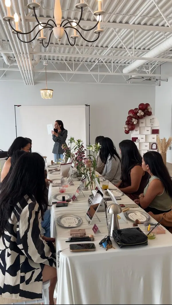 A host demonstrates a technique with a brush and mirror at the head of the table while guests follow along from their seats.