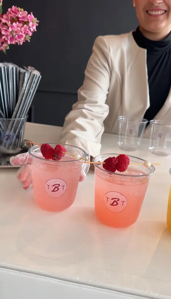 Two pink raspberry mocktails in cups printed with The Beauty Table monogram, each garnished with raspberries on a pearl pick.