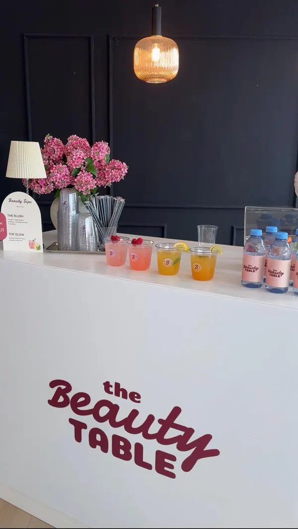 The Beauty Sips mocktail bar: pink and citrus drinks, a pink hydrangea bouquet and branded water bottles on a white counter printed with The Beauty Table logo.