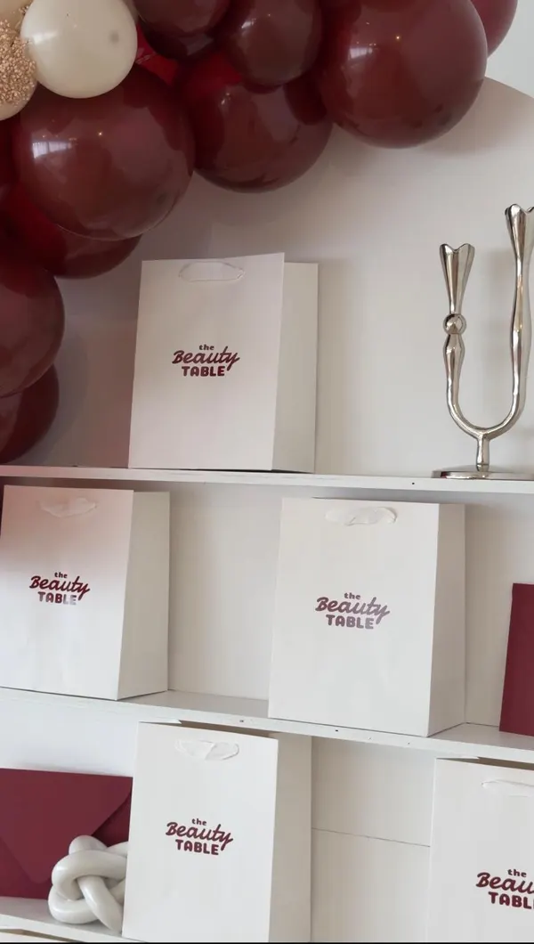 White take-home gift bags with The Beauty Table logo arranged on shelves beneath a burgundy and cream balloon garland.