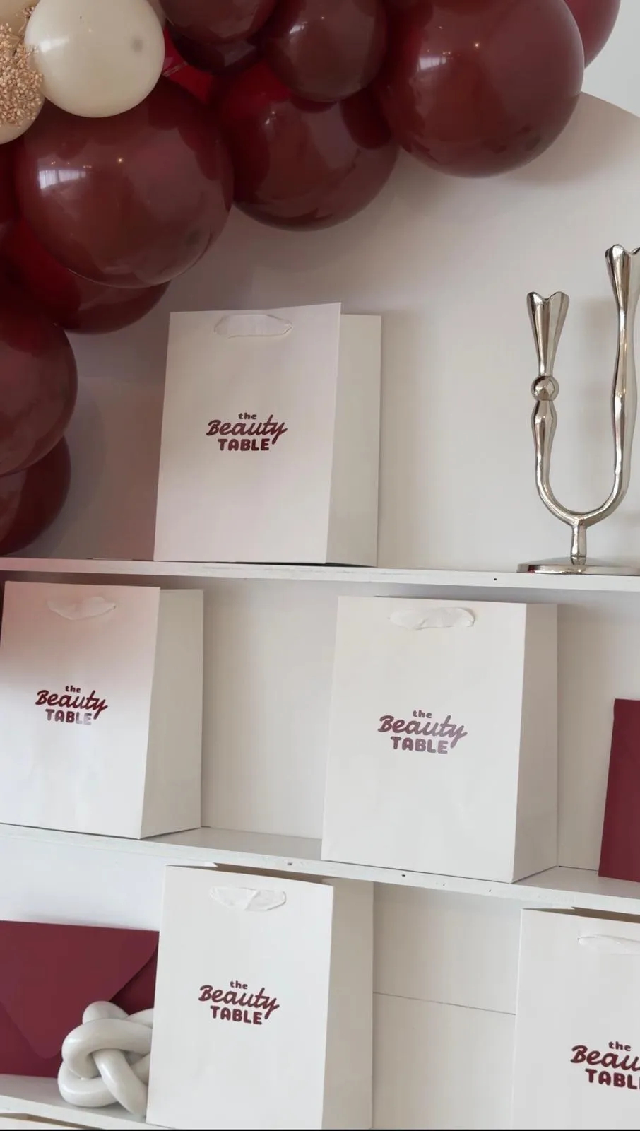 White take-home gift bags with The Beauty Table logo arranged on shelves beneath a burgundy and cream balloon garland.