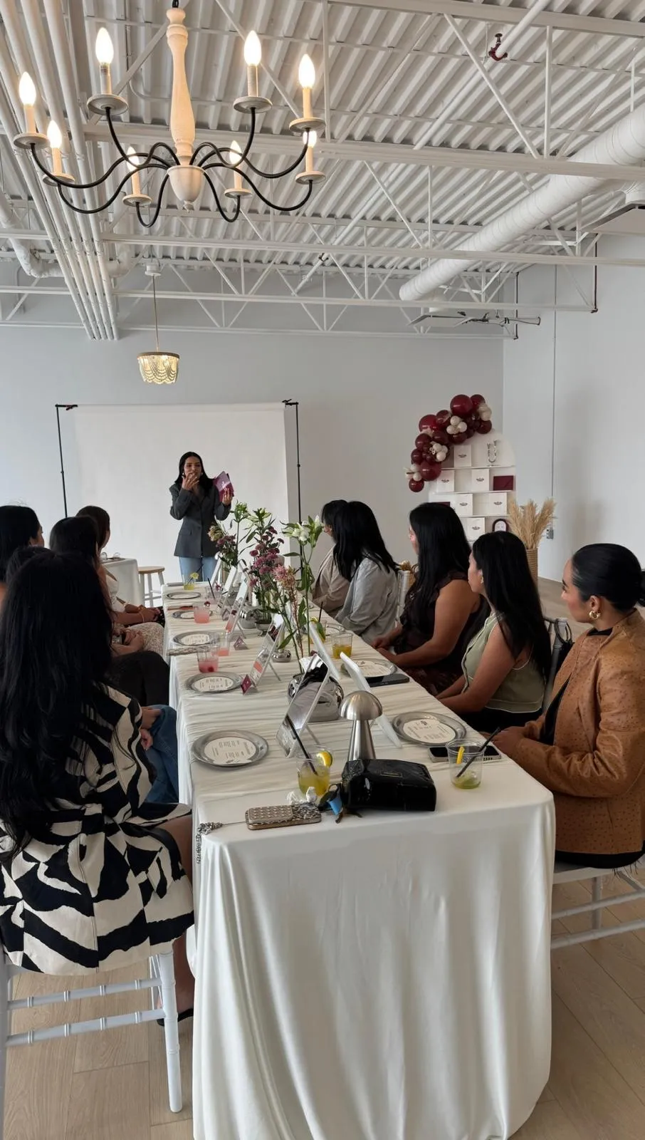 Guests seated along a long white table set with flowers and mirrors, listening as a host speaks, with a burgundy balloon garland and gift wall behind them.
