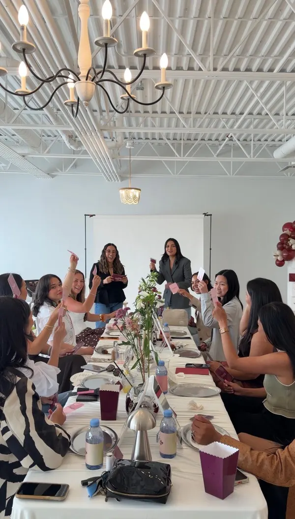 Guests around the long table laugh and hold up game cards while both hosts lead the group from the head of the table.